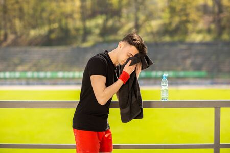 Young sporty man resting and wiping his sweat with a towel after workout sport exercises outdoors on the background of green grass.の写真素材