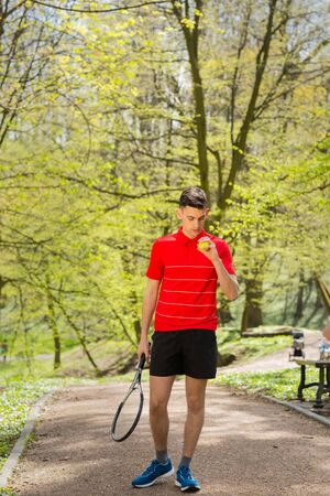 The man in a red t-shirt pose with a tennis racket and a ball on the background of green park. Sport concept.の写真素材
