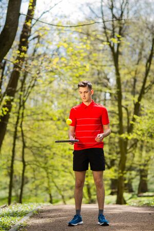 The man in a red t-shirt pose with a tennis racket and a ball on the background of green park. Sport concept.の写真素材