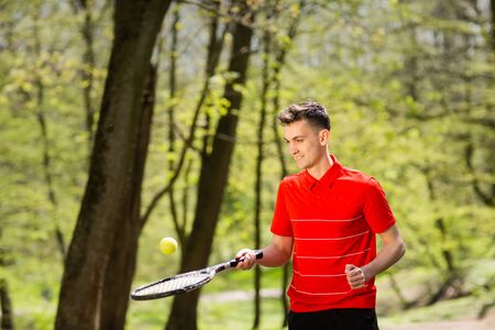 The man in a red t-shirt pose with a tennis racket and a ball on the background of green park. Sport...