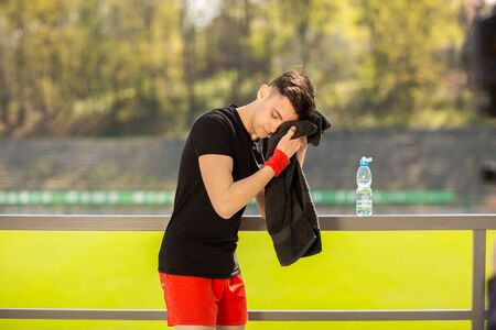 Young sporty man resting and wiping his sweat with a towel after workout sport exercises outdoors on the background of green grass.の写真素材