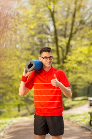 Young sports man in red t-shirt posing in a spring park with a blue yoga mat. His hands shows class.の写真素材