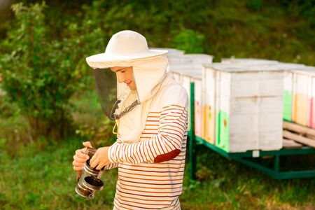 A beekeeper in a protective cap prepares a tool for smoking bees from a hive in the apiary. Smoke for bees, equipment on apiary.の写真素材