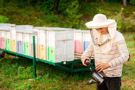 A beekeeper in a protective cap prepares a tool for smoking bees from a hive in the apiary. Smoke for bees, equipment on apiary.の写真素材