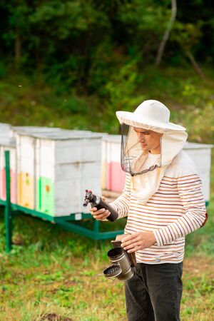 A beekeeper in a protective cap prepares a tool for smoking bees from a hive in the apiary. Smoke for bees, equipment on apiary.の写真素材