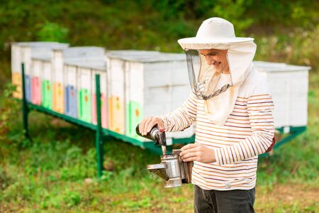 A beekeeper in a protective cap prepares a tool for smoking bees from a hive in the apiary. Smoke for bees, equipment on apiary.の写真素材