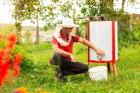 A beekeeper in a protective cap crouched near the honey extractor. He pours fresh honey into the white bucket.の写真素材