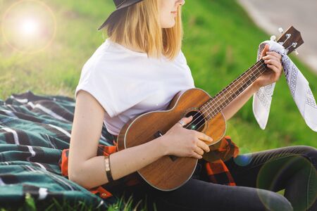 Portrait of a young beautiful woman in a black hat. Girl sitting on the grass and playing guitar.