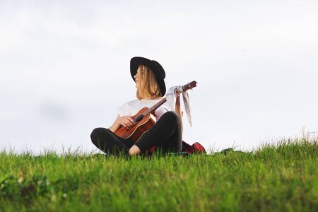 Outdoor portrait of a young beautiful woman in black hat, playing guitar. Space for text.