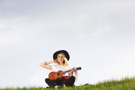 Outdoor portrait of a young beautiful woman in black hat, playing guitar. Space for text.の写真素材