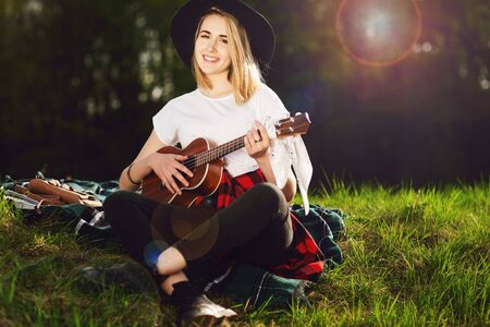 Portrait of a young beautiful woman in a black hat. Girl sitting on the grass and playing guitar.の写真素材