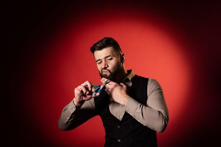 Bearded man in vest on red background. Men cut their beard with hairdressing scissors.