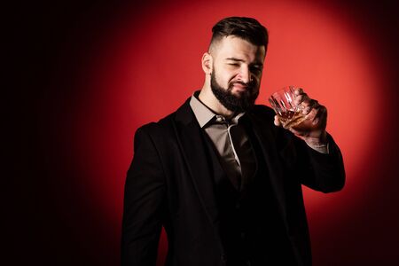 Handsome bearded man in suit with stylish beard holding a glass of whiskey and going to drink it, on red background.の写真素材