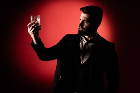 Handsome bearded man in suit with stylish beard holding a glass of whiskey and looking at him, on red background. Copy space.の写真素材