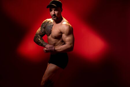 Muscular and strong man in typical bodybuilding pose. Bodybuilder with tattoo in cap on red background. Side view, copy space.の写真素材