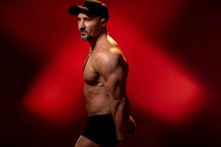 Muscular and strong man in typical bodybuilding pose. Bodybuilder with tattoo in cap on red background. Side view, copy space.の写真素材