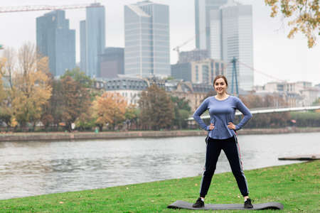 Sporty young woman in sportswear doing stretching exercises on the yoga mat near the river. Behind the panorama of the big city. Active lifestyle.の写真素材