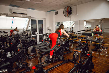 Attractive woman in a red sports suit in gym, riding on speed stationary bike. Rear view. Reflection in the mirror. Healthy lifestyle.の写真素材