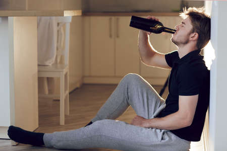 Side view, drunk young man with bottle of wine sitting on the floor in kitchen. Drinking alone. Male...