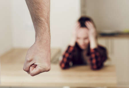 Male fist close up in focus. On a blurred background, a woman is sitting at a table, clasping her...