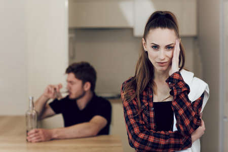 Drinking problem drunk man in a young family. Upset woman in the kitchen holding her head on the background of a drunk man. The man behind sits at the table and drinks alcoholの写真素材