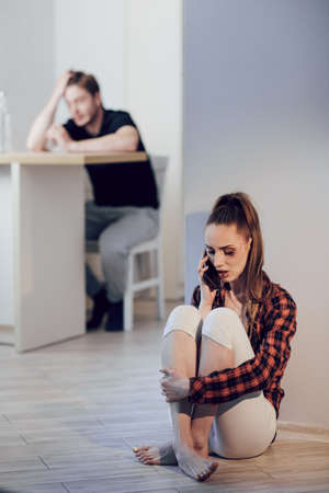 A young scared woman with bruises on her face hides from her husband and calls the police. Drunk man drinking alcohol in the kitchen. The concept of domestic violence and alcoholismの写真素材