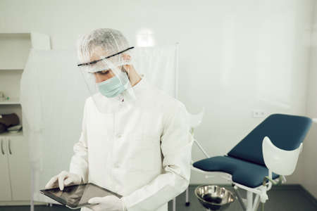 A male gynecologist with tablet in hands, in a protective suit prepares to receive patients during quarantine of coronavirus. Physician in personal protective equipment during a pandemicの写真素材