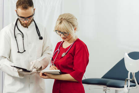 Male and female medical doctors using a tablet and write something and a notebook in a hospital office. Doctors in uniform, man and woman in a gynecological officeの写真素材