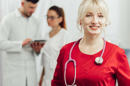 Close-up portrait of a smiling female doctor in a red uniform with a stethoscope. Young doctor smiling in posing at camera, behind her on the background two other doctorsの写真素材