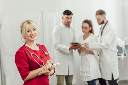 Young doctor woman in red uniform with a notebook in office. Woman looking at camera. In the background, a team of three doctors talkingの写真素材