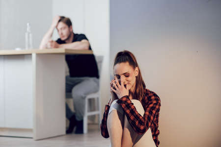 Young scared woman with bruises on face complains to girlfriend on cell phone sitting on the floor in the kitchen.の写真素材
