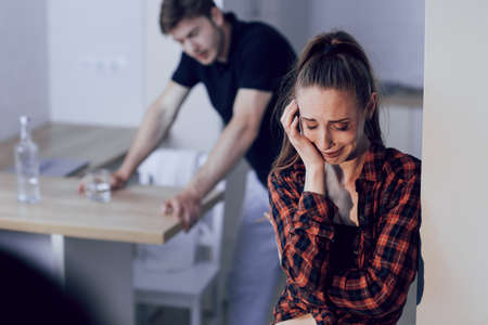 Scandal in a young family, problems of domestic violence. The frightened woman with bruises on her face is crying, the man is approaching the woman from behindの写真素材