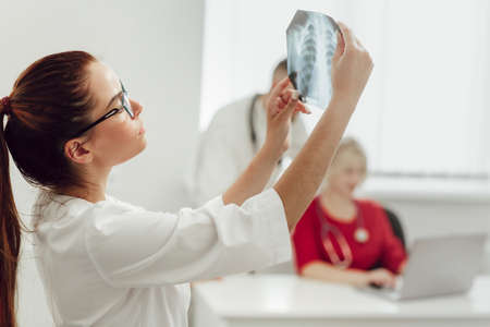 Young female doctor in hospital looking at x-ray. The other two doctors communicate in the background. Medicine and treatment conceptの写真素材