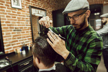 Haircut in the barbershop. Close up view of young bearded man getting haircut while sitting in chair at barbershop. Hairdresser cuts hair with scissors and a combの写真素材