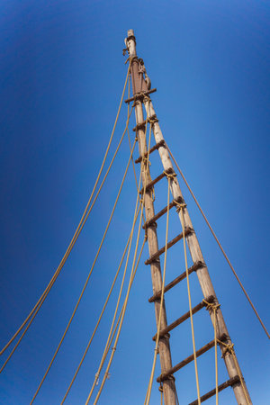 Gazing up at a tall wooden mast adorned with ropes set against a vivid blue skyの写真素材