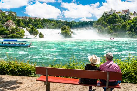 Neuhausen am Rheinfall, Switzerland - 23 July 2019. Waterfall on the river Rhine in the city of Neuhausen am Rheinfall in Switzerland.A Senior couple sitting on a bench and admiring a waterfall.のeditorial素材