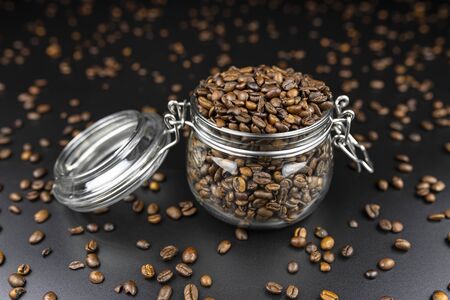 Fresh, roasted coffee beans in a open, transparent jar standing on a black table with lots of coffee scattered around.の写真素材