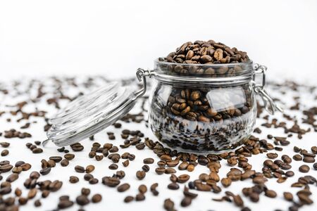 Fresh, roasted coffee beans in a open, transparent jar standing on a white table with lots of coffee scattered around, white background from the back.の写真素材