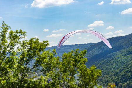 A man flying a white paraglider over the forest and the river on a beautiful sunny day.の写真素材