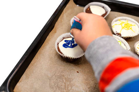 A child squeezes colored frosting from a tube onto chocolate brown cupcakes covered with white frosting with colorful decorations, isolated on white.の写真素材