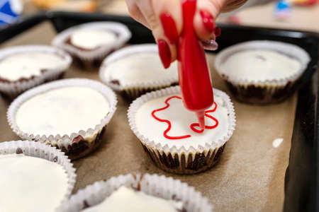 A woman squeezes colored frosting from a tube onto chocolate brown cupcakes covered with white...