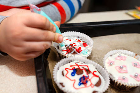A child squeezes colored frosting from a tube onto chocolate brown cupcakes covered with white...