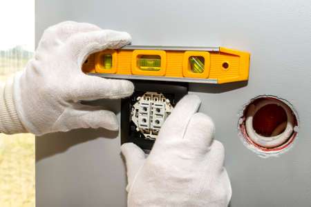 An electrician mounts the black frame of the roller shutter switch up and down with white gloves in the room by the window using a spirit level.の写真素材
