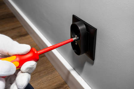 An electrician uses a red screwdriver to screw a black double grounded electrical socket into the...