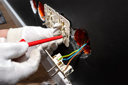 An electrician uses a red screwdriver to screw a double grounded electrical socket in a row to the electrical box in the black wall of the room.の写真素材