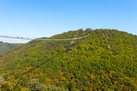 A hanging wooden bridge with steel ropes seen from the side against a blue sky, visible tourists on...