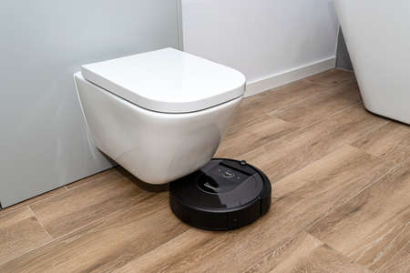 A modern robotic vacuum cleaner cleans the ceramic tiles in the bathroom next to the toilet, an...