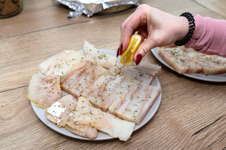 A woman squeezes lemon on a fresh cod fillet cut into squares, sprinkled with lovage.