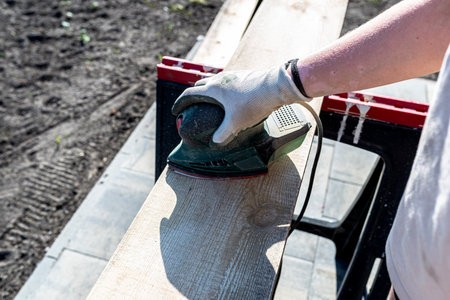 A woman sands a board with a small orbital sander placed on trestles in the garden.