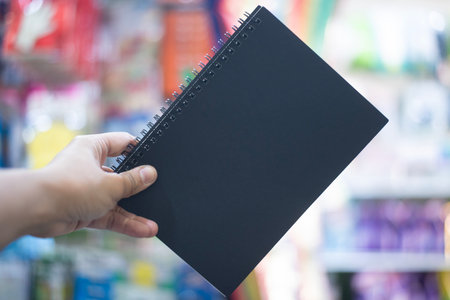 A person's hand is holding up a blank, spiral-bound notebook with a matte black cover. The hand is positioned in the lower-left corner, and the notebook is held at a slight angle. The background is a blurry, colorful scene of what appears to be a retail store aisle with shelves filled with various products. The soft focus on the background puts all the emphasis on the notebook, which serves as a canvas for creativity, planning, or note-taking. The image is clean and simple, making it perfect for mockups or concepts related to stationery, education, and creative workの写真素材